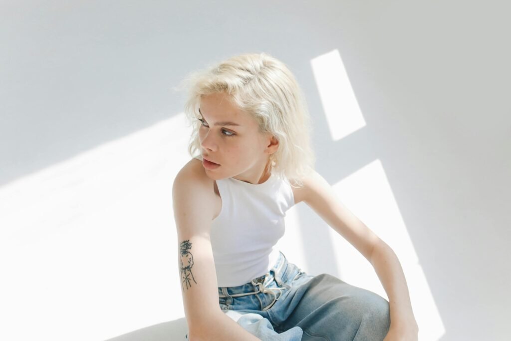 Young adult in sleeveless top sitting indoors, bathed in bright sunlight, with a contemplative expression.